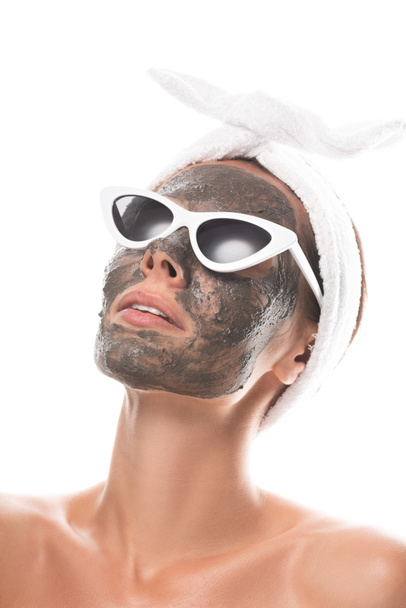 mujer joven desnuda en banda de pelo cosmético y gafas de sol con máscara de arcilla en la cara aislada en blanco
 - Foto, imagen