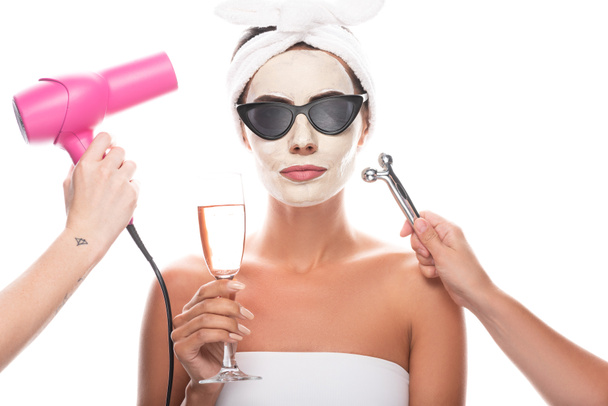 vista parcial de esteticistas sosteniendo secador de pelo y masajeador facial y mujer en gafas de sol con máscara facial sosteniendo copa de vino aislada en blanco
 - Foto, Imagen
