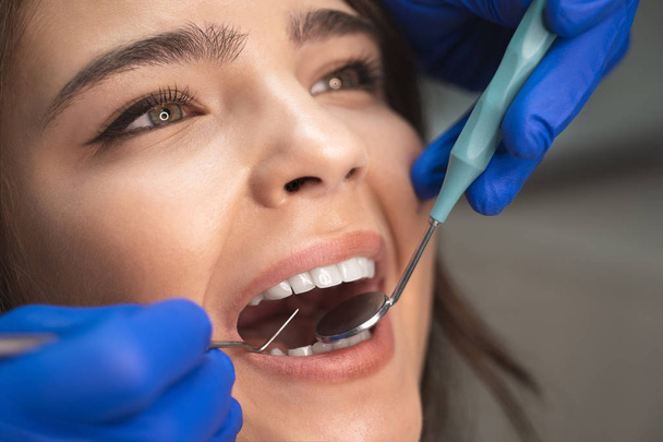 smiling beautiful happy brunette woman patient examined by dentist in blue gloves using dental mirror and scaler sitting in dental clinic close up - Фото, изображение