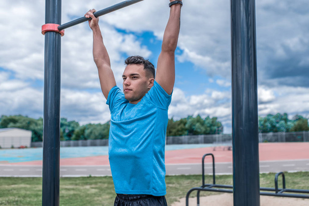 young guy, man sports athlete, in summer in city on sports ground, pulls up on bar, workout fitness, workout healthy lifestyle. Sportswear, strength and endurance, motivation. - Foto, imagen