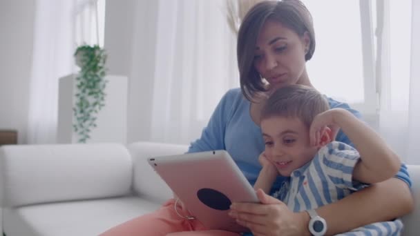 Mother and son sitting on sofa using digital tablet. Happy mom and little boy using tablet with touchscreen together watching a video. Smiling mother and cute boy playing on digital tablet - Footage, Video