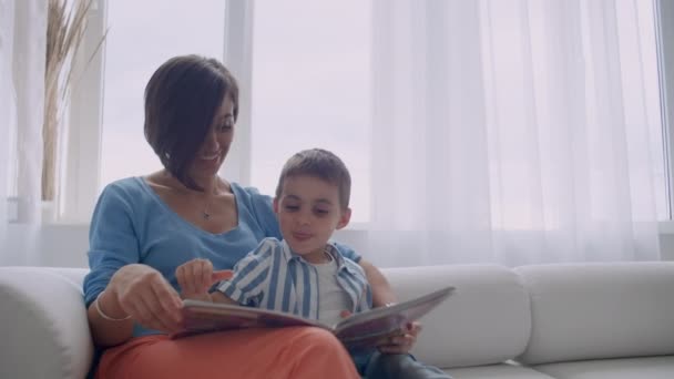 幸せな母と小さな息子は、自宅のリビングルームで一緒に朝に本を読みます。家族活動の概念 - 映像、動画