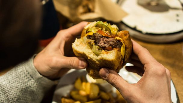 若い男の手はファーストフードレストランで噛まれたハンバーガーを保持しています - 写真・画像