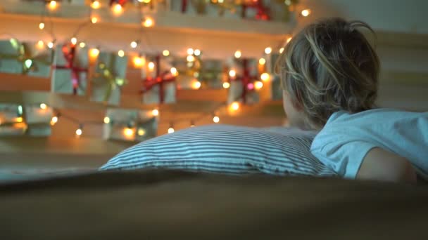 Slowmotion strzał małego chłopca budzi się i widzi Kalendarz adwentowy wiszące na łóżku rozjaśnić z lampami świątecznymi. Przygotowanie do Bożego Narodzenia i nowego roku koncepcji. Koncepcja kalendarza adwentowego - Materiał filmowy, wideo