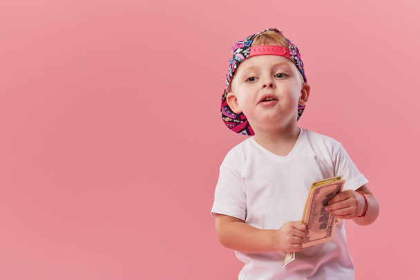 Portrait of cheerful little toddler boy in cap holding dollars money on pink background - Fotó, kép