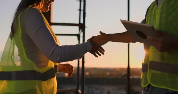 Будівельники чоловік з планшетом і жінка в білих шоломах тремтять руками під час заходу сонця, стоячи на даху будівлі
. - Кадри, відео