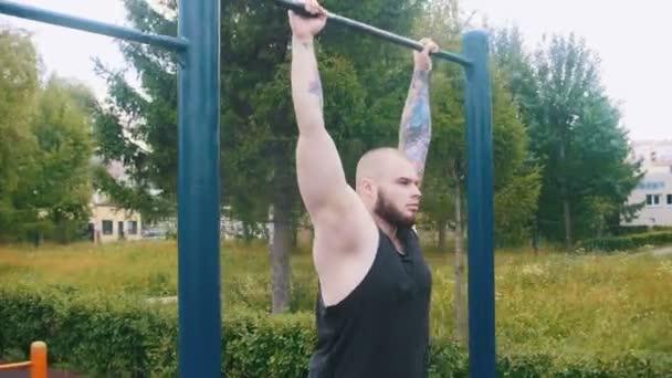 A big bearded man with tattoos hanging on the bar on a sports ground outdoors - Кадры, видео
