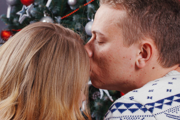 Couple in love sitting next to a Christmas tree, wearing warm sweaters, hugging and looking away from the camera towards the tree. - Photo, Image