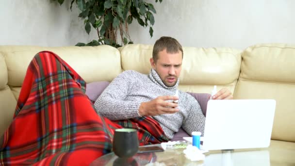 άνθρωπος με ένα κρύο κάθεται στον καναπέ σε ένα πουλόβερ και ένα καρό καλώντας το γιατρό του σε ένα φορητό υπολογιστή μέσω βίντεο Link. - Πλάνα, βίντεο
