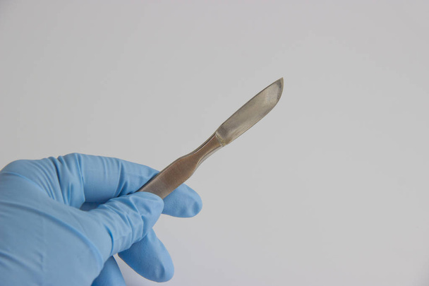 Рука хирурга в синей медицинской перчатке со скальпелем на белом фоне
 - Фото, изображение