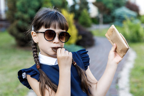 Linda chica, años de escuela vestida con uniforme escolar con placer comiendo chocolate negro en la calle en el parque
 - Foto, Imagen
