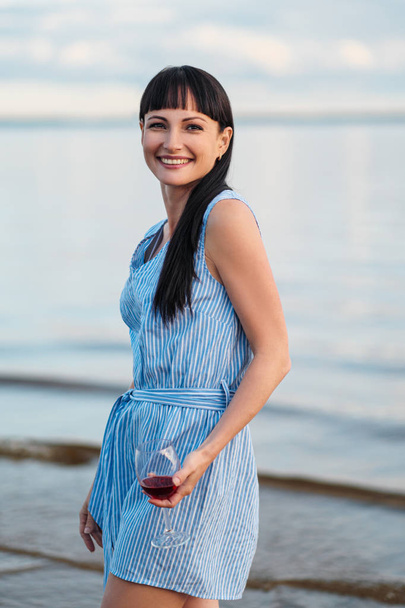 A young attractive brunette woman, in a blue dress, walks along the seashore. Portrait of a smiling woman on vacation with a glass of wine. - Foto, afbeelding