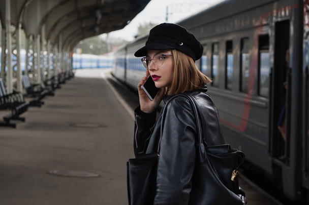 Attractive young woman millenial in black clothes and a hat at the railway station next to the train. Speaks on a mobile phone, smiles and waits for the train. - Fotó, kép