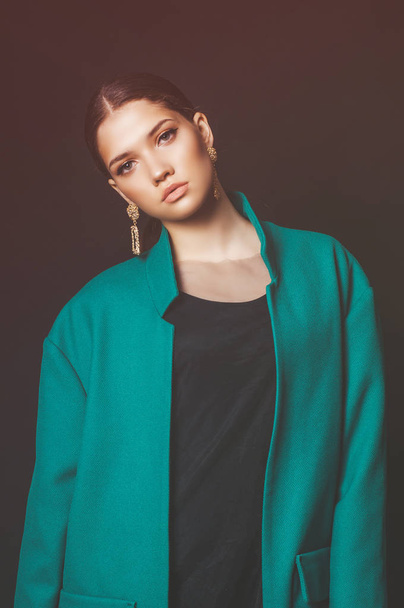 Attractive young girl model with dark hair in a trendy green jacket and jewelry on a black background in the studio - Photo, Image