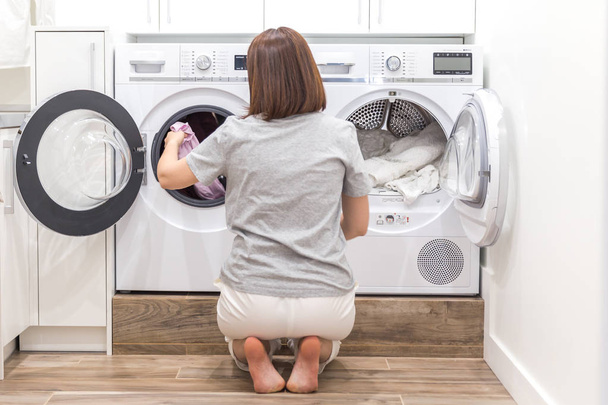 Женщина загружает грязную одежду в стиральную машину для стирки в современной подсобке
 - Фото, изображение