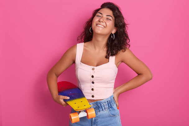 Magnificent girl with charming smile in casual fashionable clothes posing with skateboard in hands. Studio portrait of beautiful female posing isolated over pink background. People emotion concept. - Фото, изображение