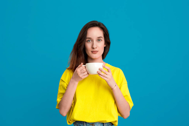 Bella donna bruna in una t-shirt gialla con una tazza bianca con una bevanda in piedi su sfondo blu
. - Foto, immagini