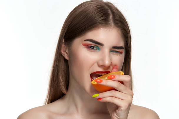 顔の近くにオレンジの半分を噛む明るい色のメイクアップを持つ美しい若い女性のクローズアップ肖像画. - 写真・画像