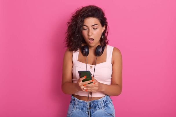 Image of impressed shocked young lady opening her mouth widely with shock, looking at device screen attentively, watching impressive content, wearing pink top and jeans. Technologies concept. - Foto, imagen