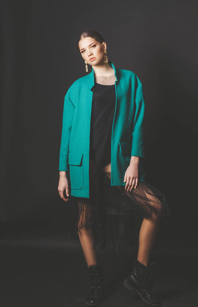 Attractive young girl model with dark hair in a trendy green jacket and jewelry on a black background in the studio - Foto, immagini