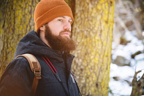 Close-up portrait of a bearded young man in a hat and a backpack in the coniferous forest - Fotó, kép