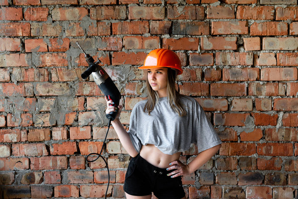 レンガの壁の背景にドリルを保持するオレンジ色のヘルメットで作業形態の女性. - 写真・画像