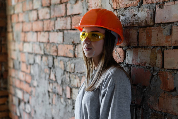 encantadora chica constructora sobre un fondo de pared de ladrillo en un casco protector naranja y gafas amarillas
. - Foto, imagen