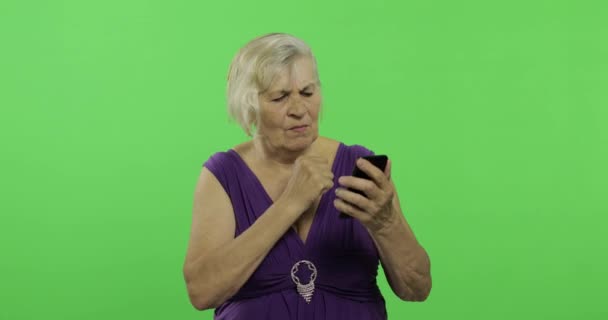 An elderly woman works on a smartphone. Old grandmother smiles. Chroma key - Footage, Video
