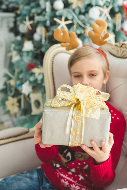 little boy holding gift box while sitting in armchair at decorative room with christmas tree on background - Φωτογραφία, εικόνα