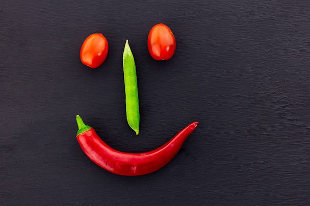 face with a smile of vegetables pepper green nose, a red pod smile and a pair of tomato eyes on a black background - Photo, image