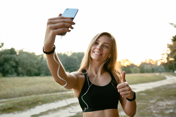 Smiling sporty girl showing a thumb up while taking a selfie outdoors - Fotoğraf, Görsel
