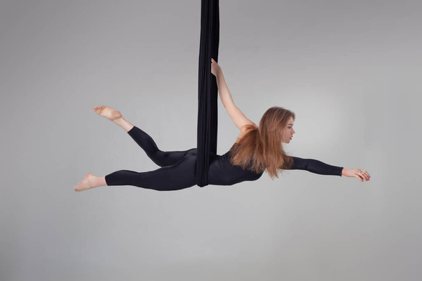 Beautiful girl in a black sport suit is performing an acrobatic elements in a studio. - Фото, изображение