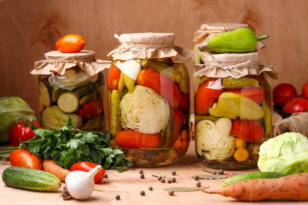 Verschiedene eingelegtes Gemüse in Gläsern: Gurken, Tomaten, Kohl, Zucchini und Paprika mit Knoblauch, Dill und Lorbeerblättern in Gläsern auf Holzgrund, horizontales Foto - Foto, Bild
