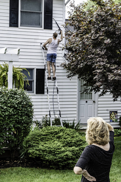 hombre en pantalones cortos en escalera poder lavado blanco casa de dos pisos, mientras que la mujer mira cerca del árbol en el patio trasero ajardinado en el día de verano soleado
 - Foto, Imagen