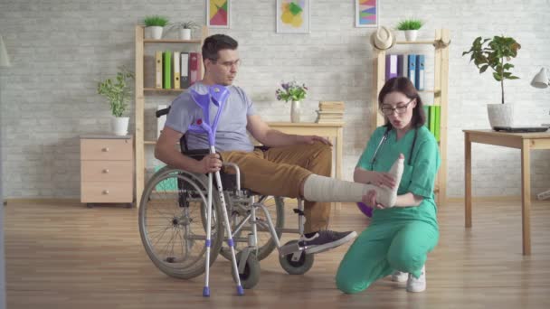 physiotherapist examines the broken leg of a man in a wheelchair after an injury - Materiał filmowy, wideo