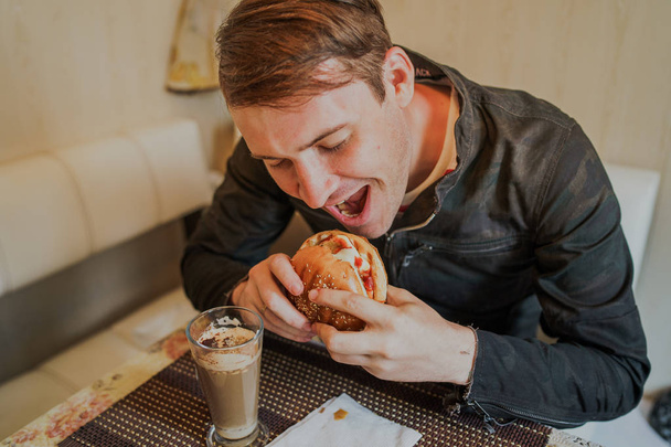 Happy man is eating fast food, hamburger. A man is sitting at the table and eating a cheeseburger - Foto, Bild