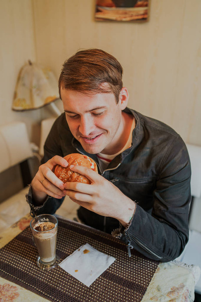 Happy man is eating fast food, hamburger. A man is sitting at the table and eating a cheeseburger - Foto, Imagem