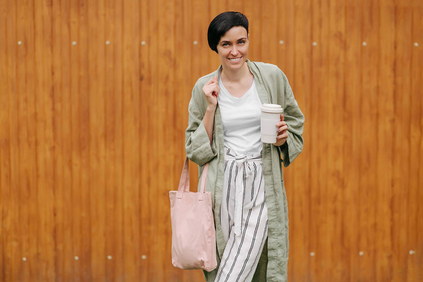 Woman with short hair holding reusable coffee cup and eco bag enjoying morning. Eco friendly concept. - Fotoğraf, Görsel