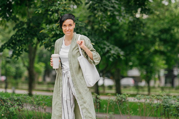 Woman with short hair holding reusable coffee cup and eco bag enjoying morning. Eco friendly concept. - Foto, Bild