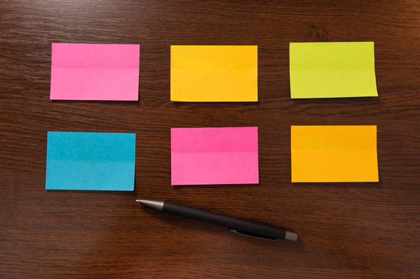 Six colorful sticky notes in two row and pen on brown wooden desk. Top view and space for text. Close up, selective focus - Photo, Image