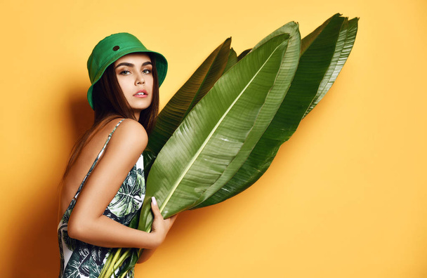 Beautiful young fashion girl with perfect skin in green hat hold tropical banana leaf in hands and covers - Foto, immagini