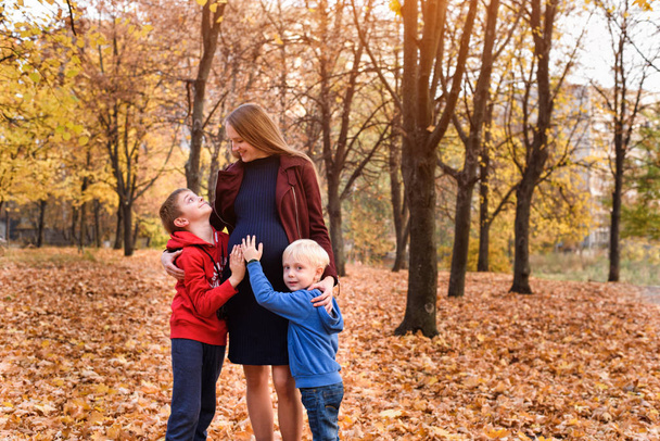 Zwei Jungen umarmen ihre schwangere Mutter. Familienkonzept. Herbstpark im Hintergrund - Foto, Bild