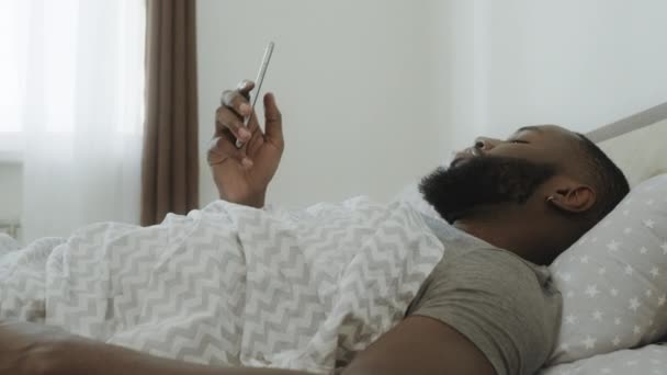 schockierter schwarzer Mann sitzt im Bett. Smartphone fällt Mann aus der Hand. - Filmmaterial, Video