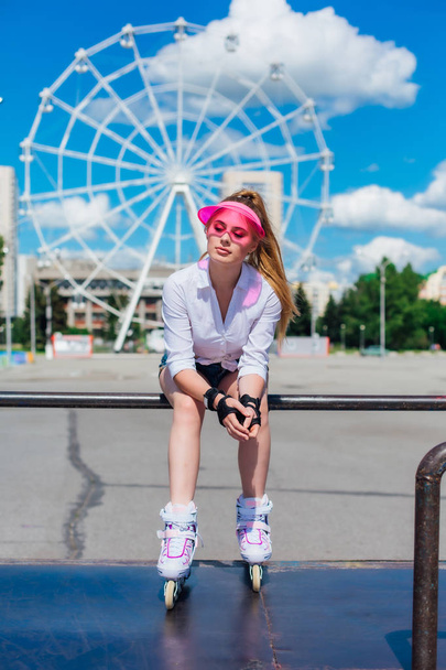 Portrait of an emotional girl in a pink cap visor wearing protective gloves and rollerblades sitting on the background of ferris wheel. - Zdjęcie, obraz