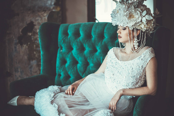 Beautiful young bride wearing white dress and wreath of flowers sits on a chair. Luxury palace. - Fotografie, Obrázek