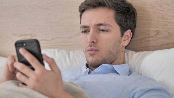 Komea väsynyt nuori mies käyttää älypuhelinta sängyssä
 - Valokuva, kuva