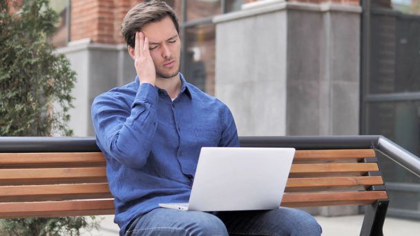 Hombre joven sentado al aire libre con dolor de cabeza usando el ordenador portátil
 - Foto, Imagen