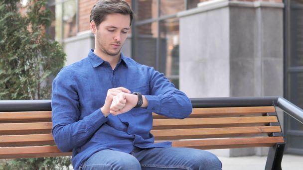 Молодий чоловік використовує смарт-годинник, на відкритому повітрі сидить на лавці
 - Фото, зображення
