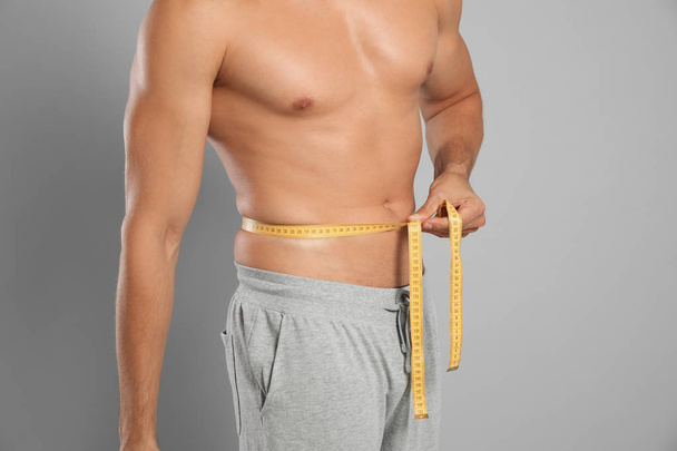 Young man with slim body using measuring tape on grey background, closeup view - Foto, Imagen