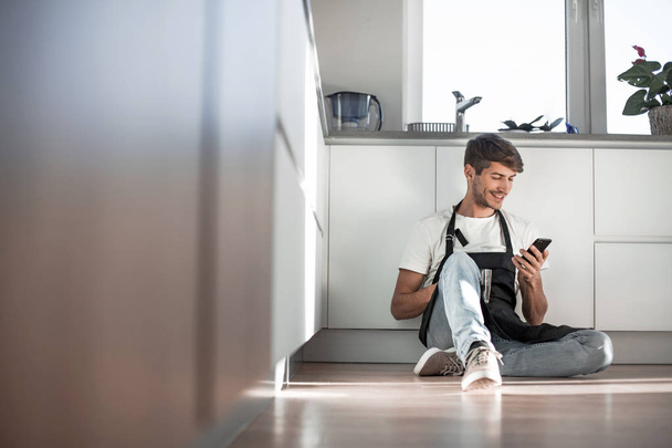 young man with smartphone sitting on kitchen floor - Foto, afbeelding